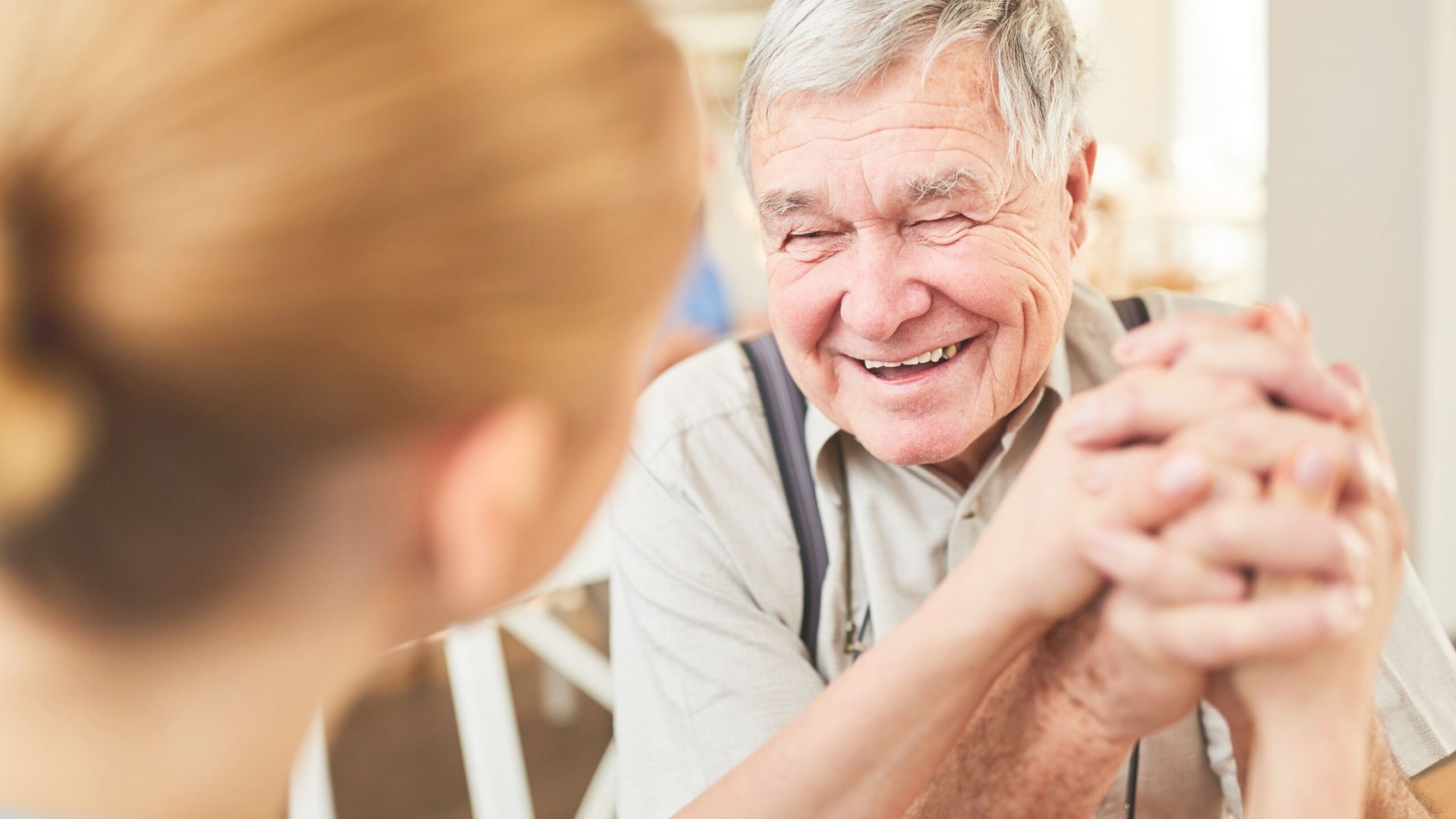 An elderly man is sitting opposite a younger woman. They are holding hands and laughing.