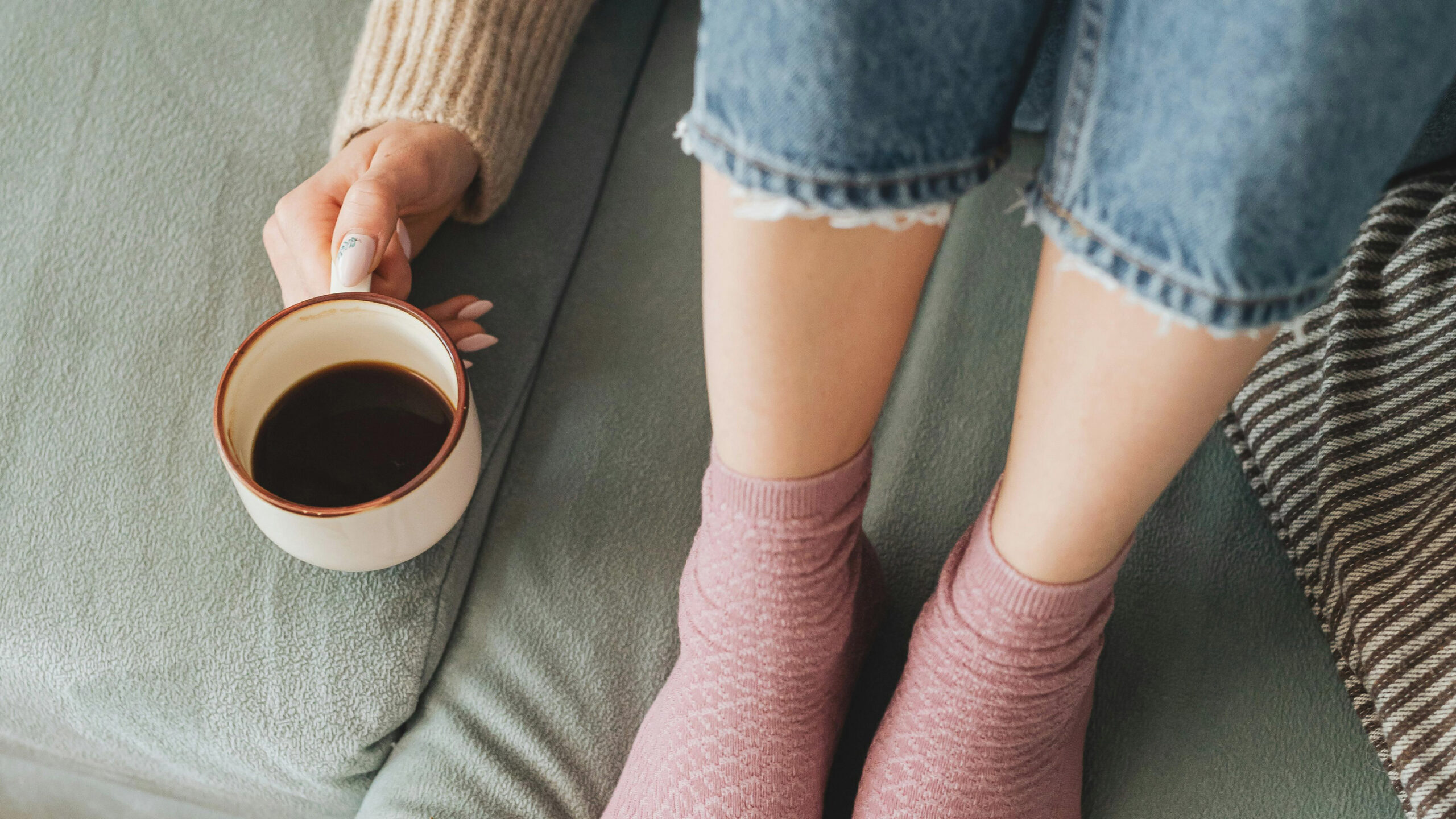 En person sitter på en soffa med en kaffekopp bredvid sig. Man ser bara fötterna på personen.