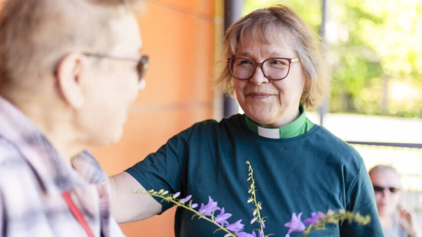 Kaksi ihmistä keskustelee ulkona. Toisella heistä on päällään diakonin vihreä virkapaita ja pitää kädessään kukkia. Toinen hymyilee ja näyttää ystävälliseltä. Taustalla näkyy vihreitä puita.
