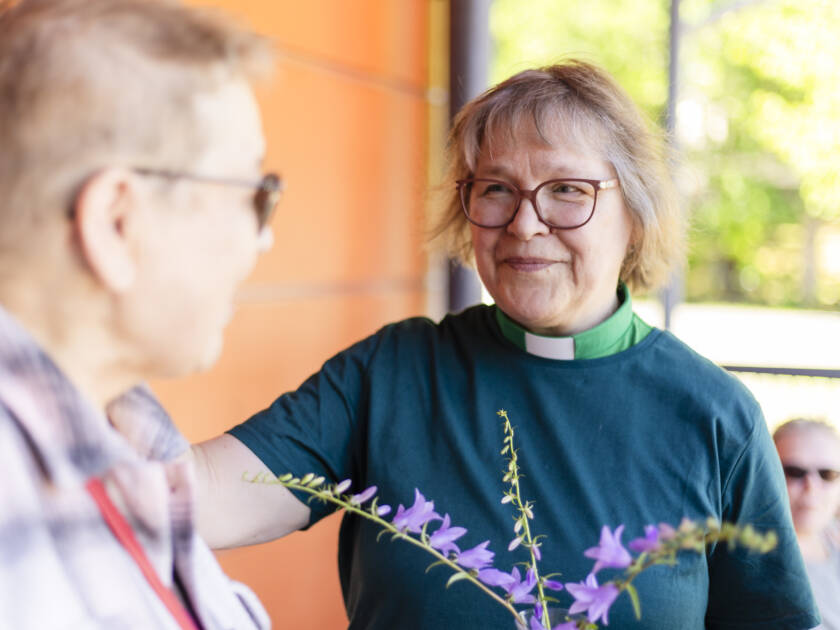 Kaksi ihmistä keskustelee ulkona. Toisella heistä on päällään diakonin vihreä virkapaita ja pitää kädessään kukkia. Toinen hymyilee ja näyttää ystävälliseltä. Taustalla näkyy vihreitä puita.