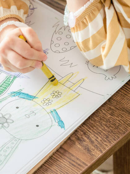 Pieni lapsi värittää värityskuvaa, joka on pääsiäisteemainen. Lapsella on keltaraidallinen paita ja hän käyttää keltaista liitua.