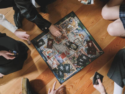 Mustiin pukeutuneet nuoret pelaavat Cluedo-peliä istuen puulattialla.