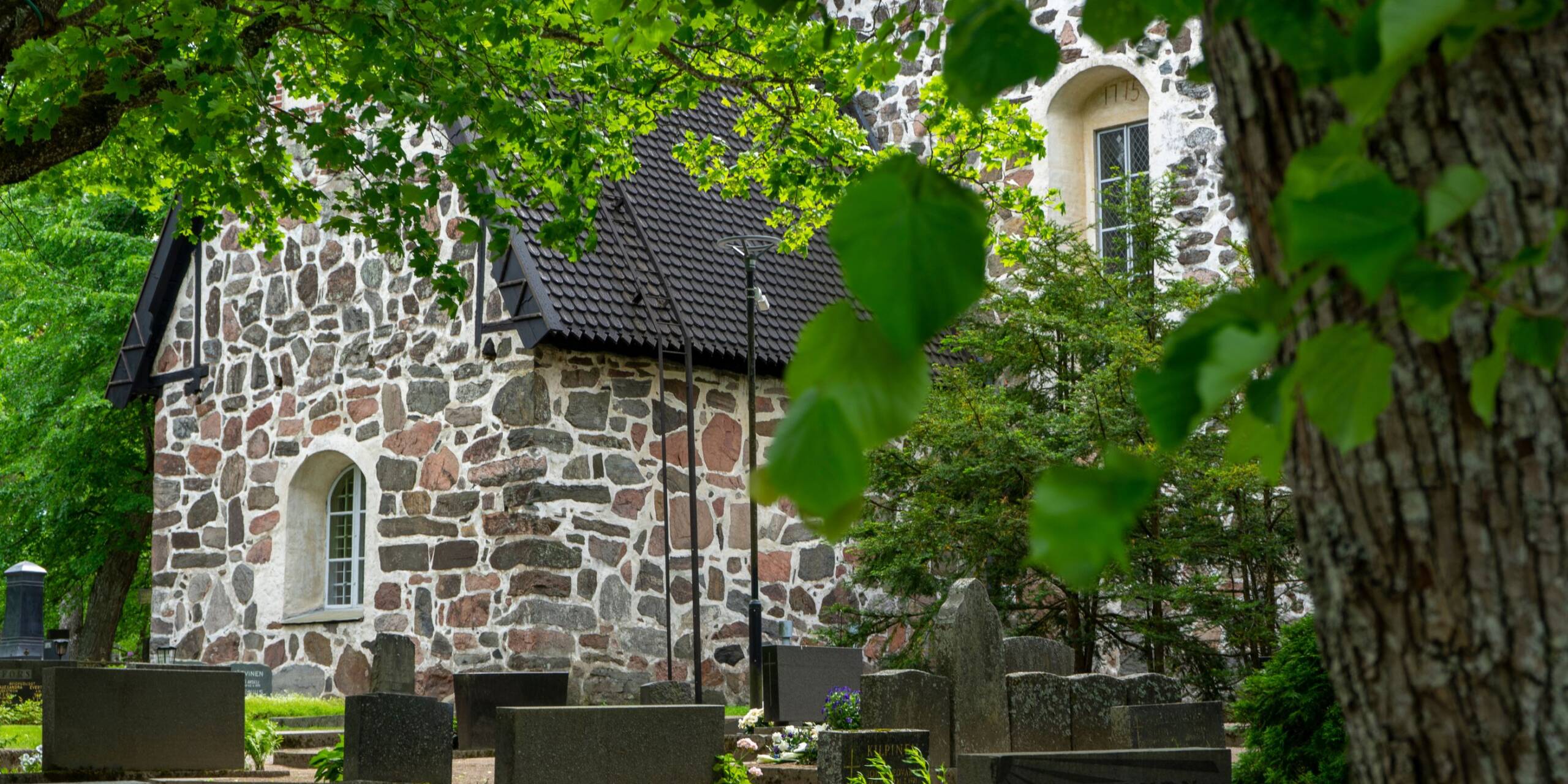 Vanha kivikirkko vehreän puutarhan keskellä, hautakiviä etualalla.