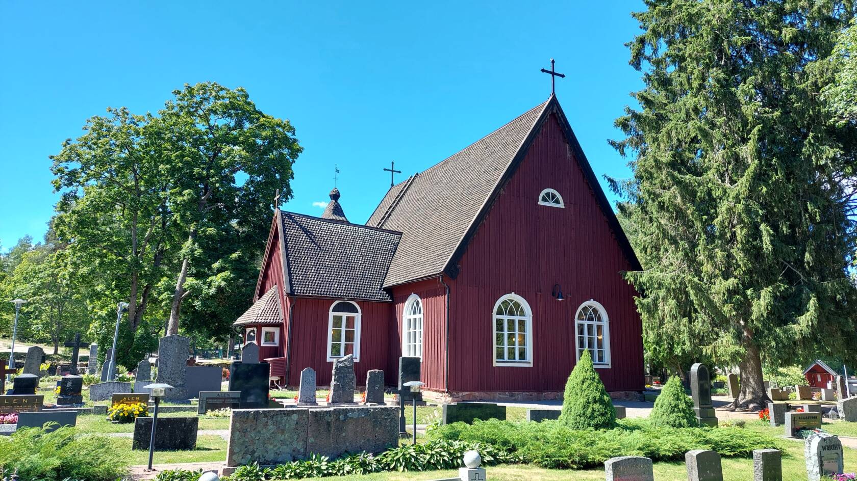 En röd kyrka under en molnfri sommarhimmel. Lövträd omger kyrkan. I förgrunden gravstenar.