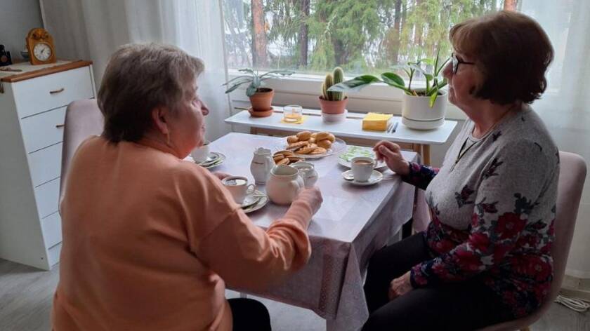 Kaksi vanhempaa naishenkilö istuu pullakahveilla keittiön pöydän ääressä ikkunan edessä.
