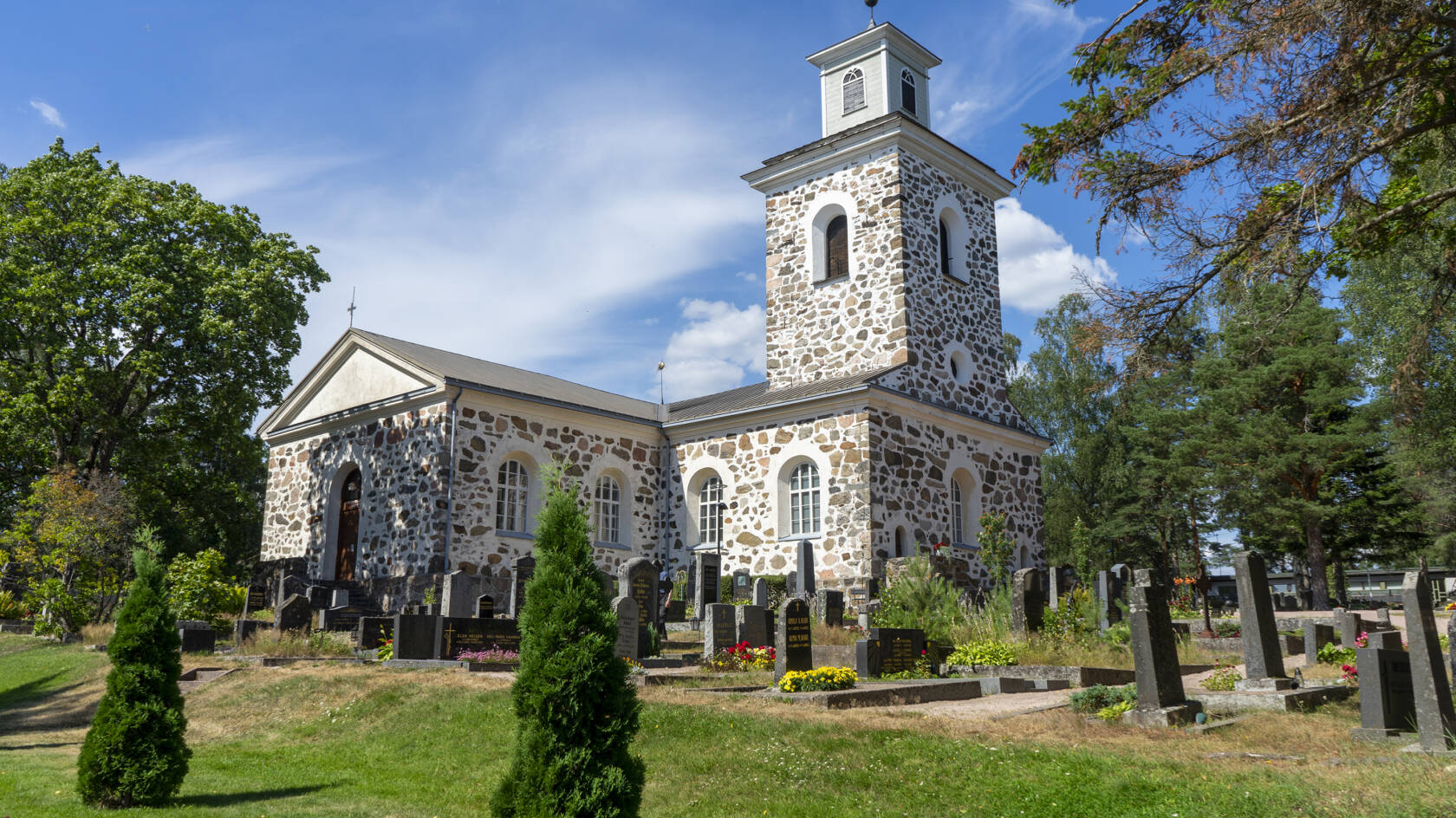 En ljus stenkyrka på sommaren med gravstenar i förgrunden.