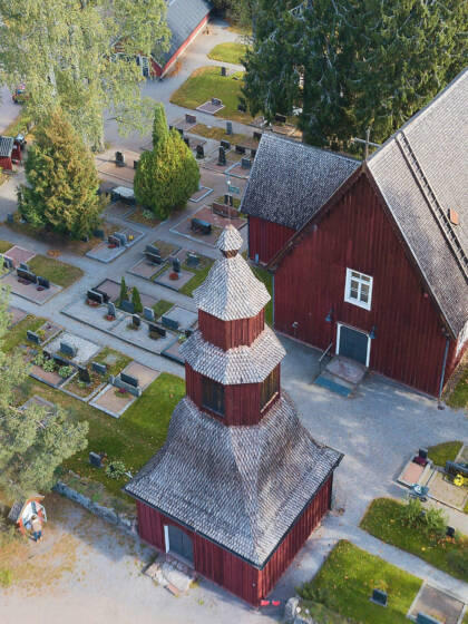 Ilmakuva punaisesta puukirkosta ja hautausmaasta, ympärillä puita ja nurmialueita.