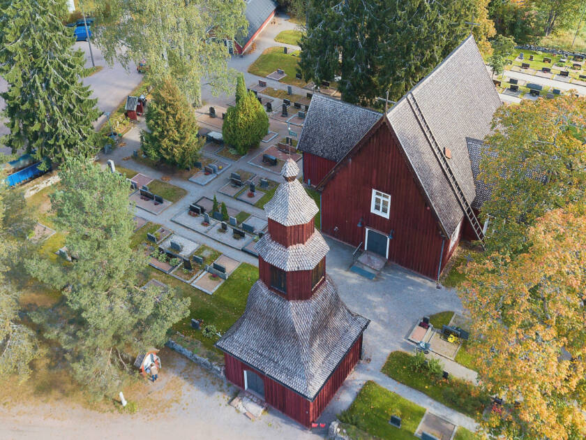 Ilmakuva punaisesta puukirkosta ja hautausmaasta, ympärillä puita ja nurmialueita.