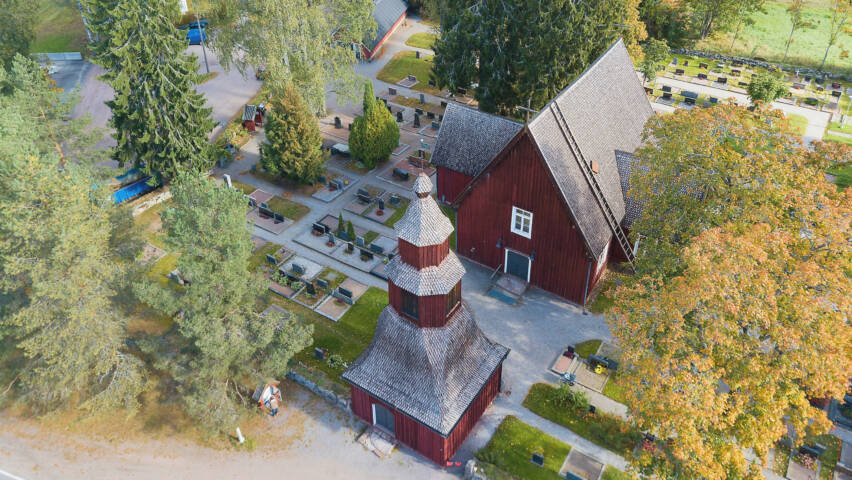 Ilmakuva punaisesta puukirkosta ja hautausmaasta, ympärillä puita ja nurmialueita.