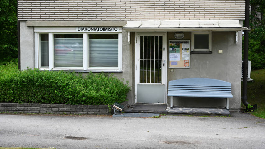 Diakoniatoimiston sisäänkäynti. Harmaa julkisivu, ovi jonka oikealla puolella sininen penkki. Oven vasemmalla puolella ikkunoita.