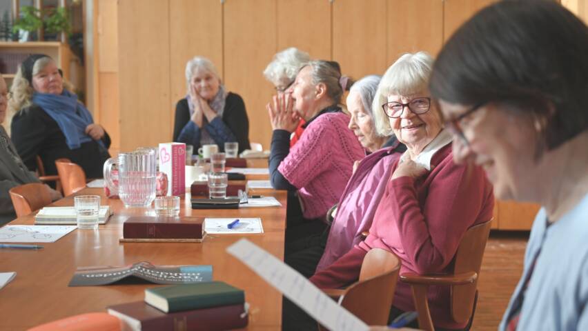 Ikäihmiset istuvat pöydän ääressä seurakuntasalissa. Etualalla ihminen lukee ja muut kuuntelevat.